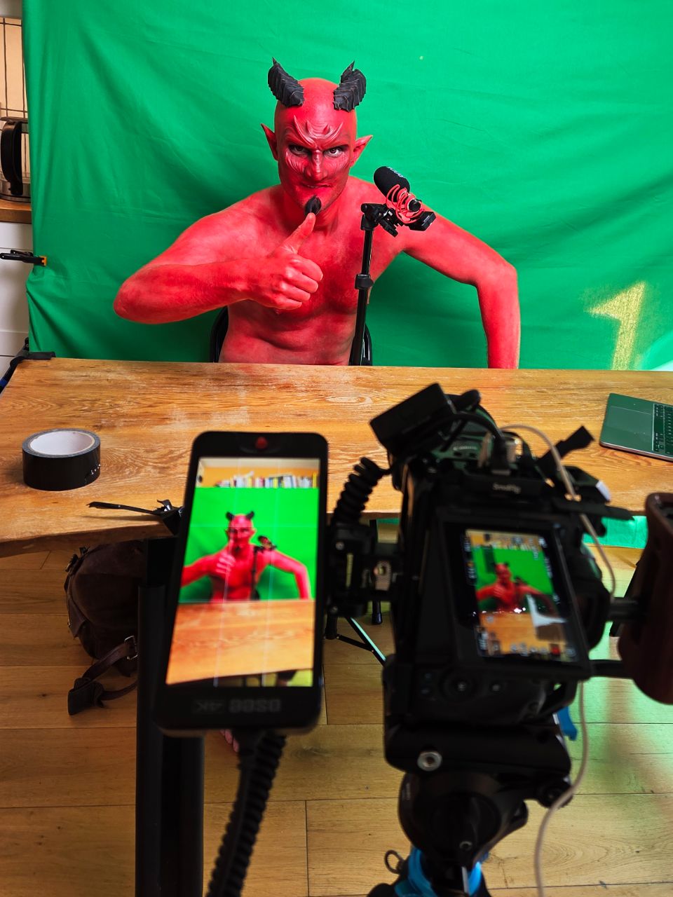 Behind the scenes of a comedy sketch shoot: a red devil character on a green screen, framed by two camera viewfinders in the foreground.